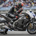 Un pilote MotoGP ajustant son casque près de sa moto sur un circuit sous une lumière naturelle du jour.